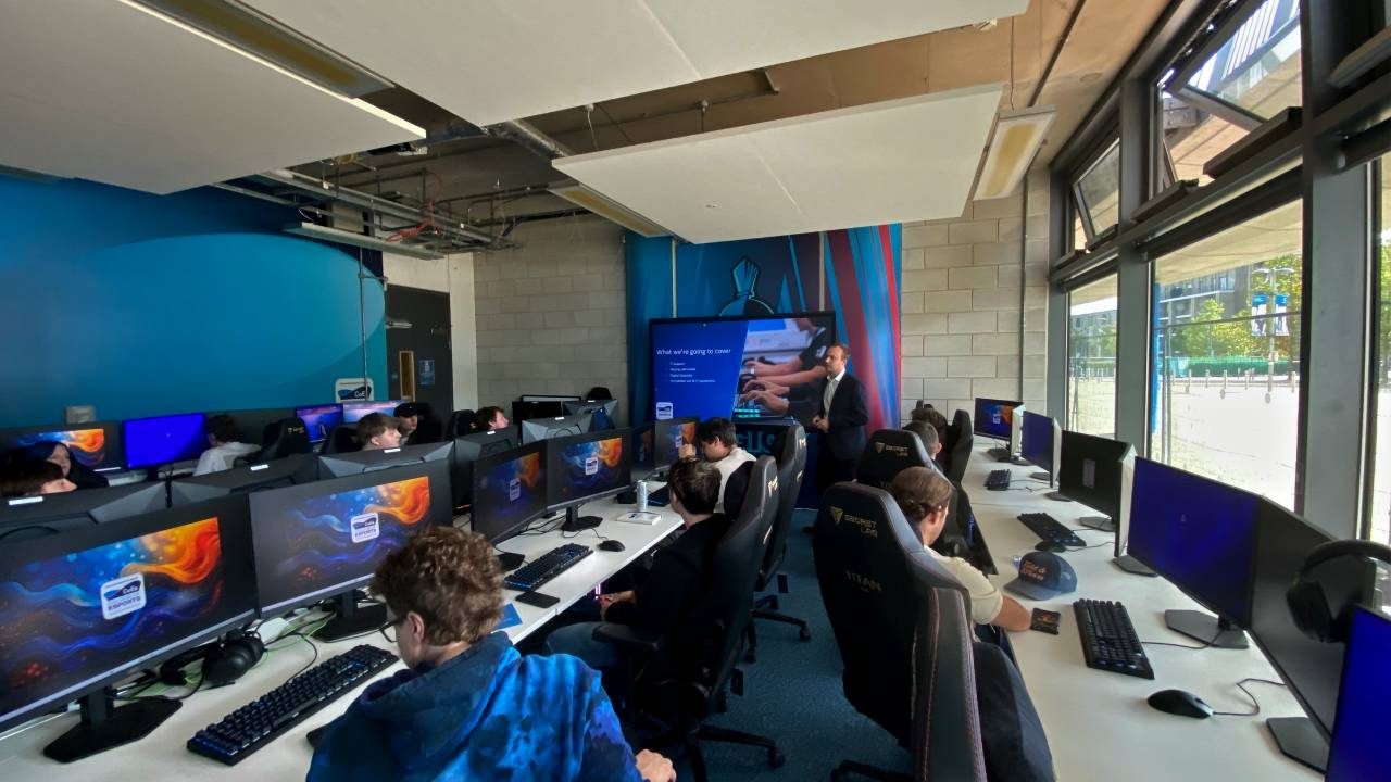 Students working on desktop computers in College of eSports modern classroom with large windows, blue and white walls, and a teacher presenting at the front near a large screen.