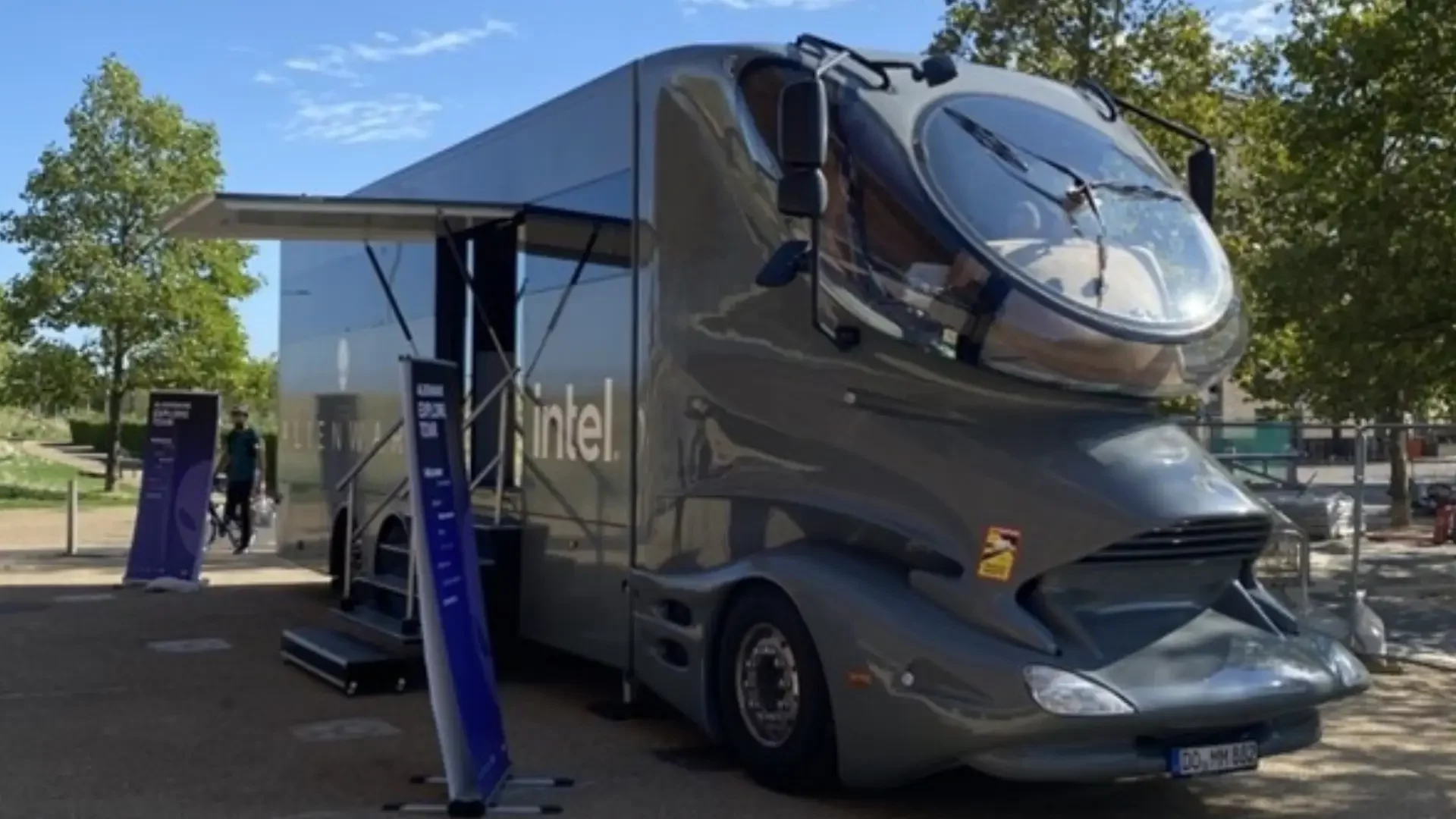 Exterior view of Dell's Alienware truck, a large grey truck, with access steps and Alienware-branded flags outside. Truck sits on brown tarmac.