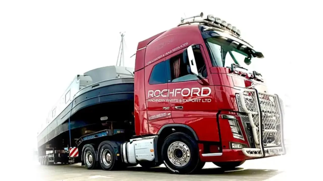 Red flatbed lorry with paint reading 'Rochford Machinery Parts & Export Ltd', viewed from close to the ground, with a boat fixed to the truck.