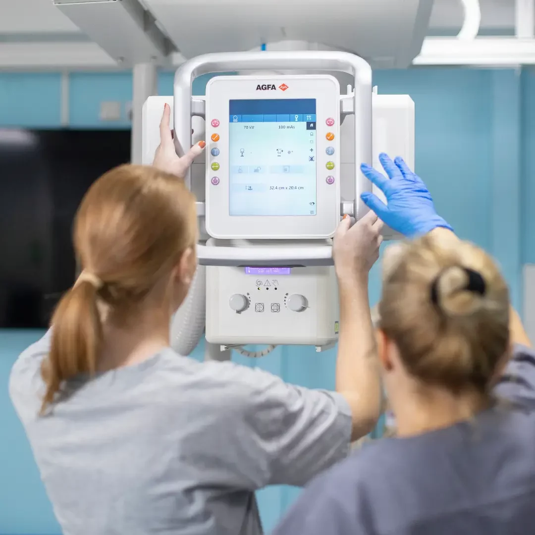 Two healthcare professionals operating an AGFA medical imaging device; one is adjusting the touchscreen control panel while wearing blue gloves, and the other is assisting, both facing the machine in a clinical setting.