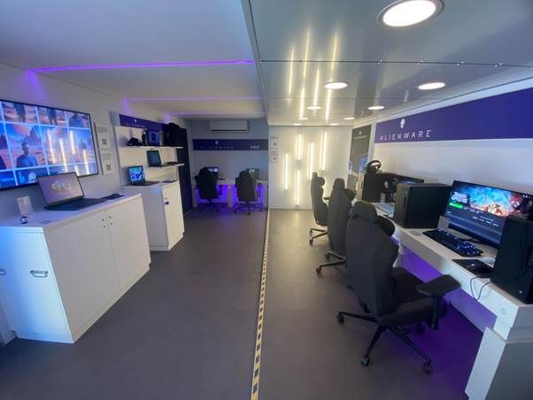 Interior of Dell Alienware truck, with multiple gaming consoles, laptops and gaming chairs plus a digital screen. Room is lit in blue and white with 'Alienware' branding.