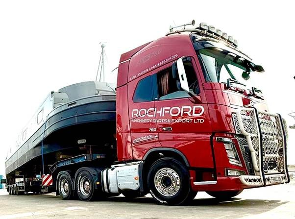 Red flatbed lorry with paint reading 'Rochford Machinery Parts & Export Ltd', viewed from close to the ground, with a boat fixed to the truck.